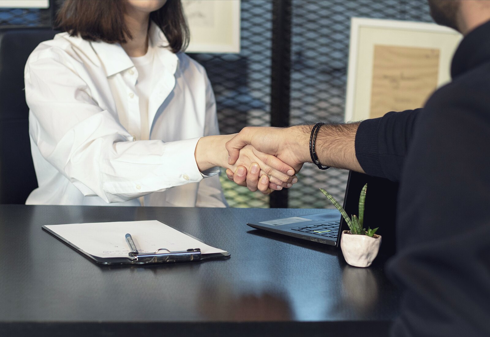 Two people shaking hands across an office desk, a signed document and pen resting on a clipboard beside them.